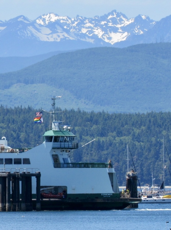 A ferry with a pride flag is docked at a pier, set against green hills and snow-capped mountains.