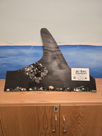 A sculpture resembling a large fin with textured and decorative elements, placed on a wooden cabinet against a blue wave-like backdrop.
