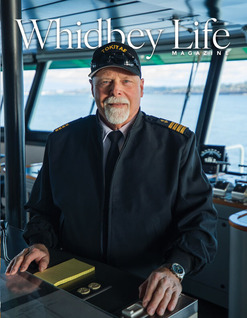 A person in a captain's uniform stands in a ferry's pilothouse with ocean views through large windows.