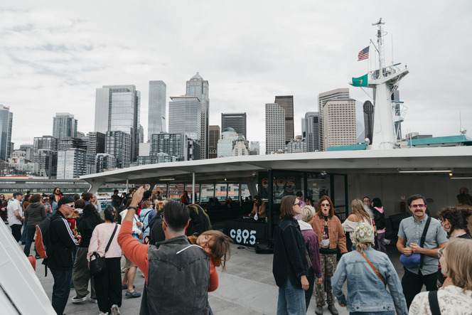 A ferry deck crowded with people, set against a city skyline of tall skyscrapers and an overcast sky.