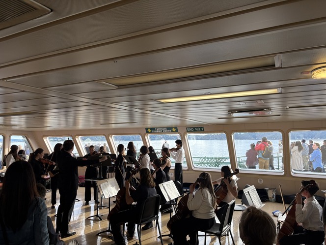 Orchestra ensemble playing inside a ferry with large windows showing the water and passengers outside.