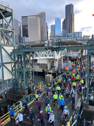 Cyclists in bright jackets disembark a ferry in front of a city skyline.