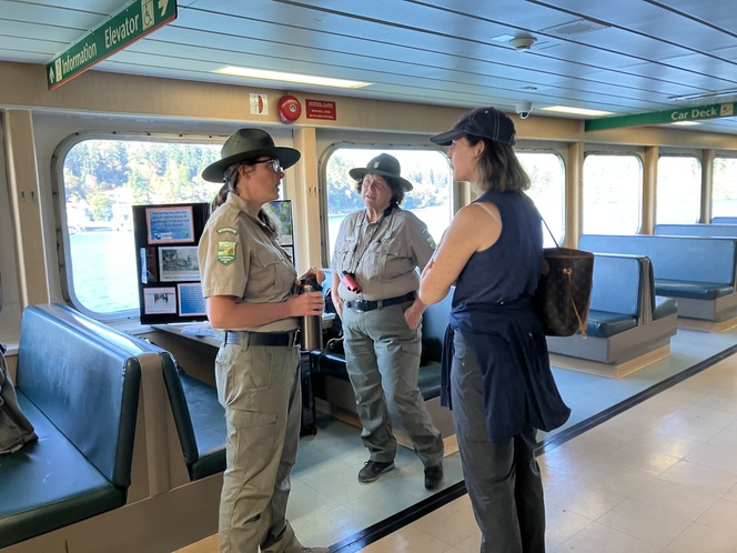 Three individuals conversing inside a ferry cabin; two are khaki-clad park rangers and one is a woman in casual attire.