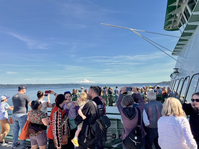 A diverse group of people on a ferry, enjoying a sunny day, with a snow-capped mountain visible in the background.