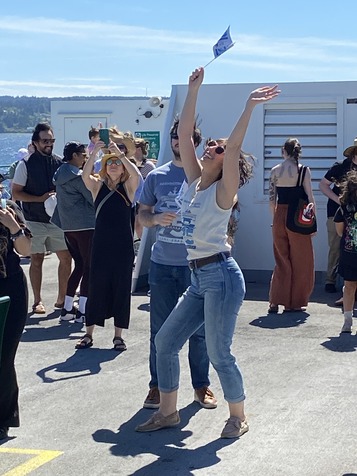 A person in sunglasses joyfully waves a small flag, standing on a sunny outdoor deck of a ferry with others in a casual, festive atmosphere.