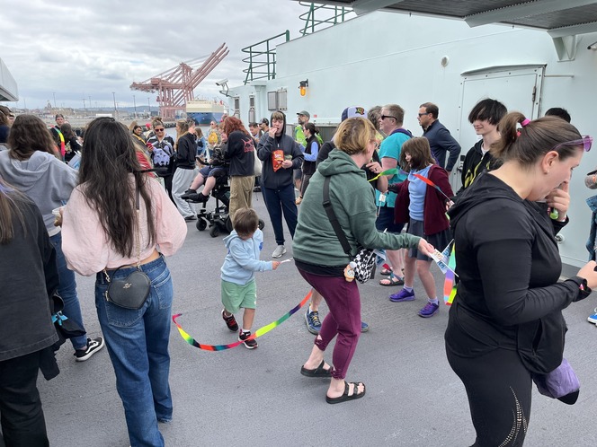 A group of people of varying ages dancing on a ferry's sun deck, some waving colorful ribbons. The scene is lively and set against an overcast sky.