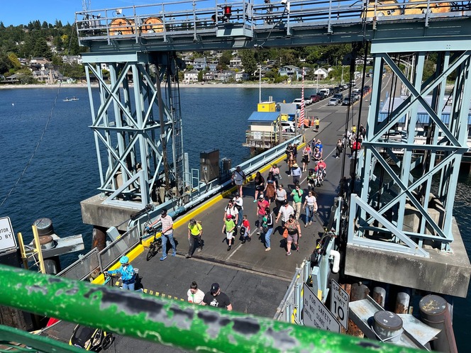 A crowded Fauntleroy terminal with a mix of pedestrians and cyclists on a pathway, surrounded by blue metal structures.