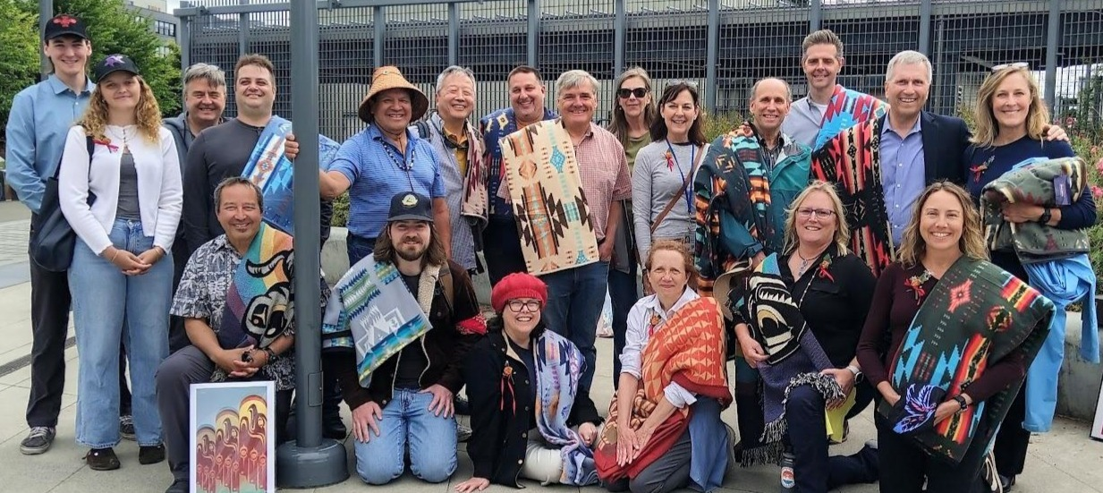 A group of 20 people posing outdoors, wearing colorful shawls and smiling, in front of a grid-like structure and greenery.