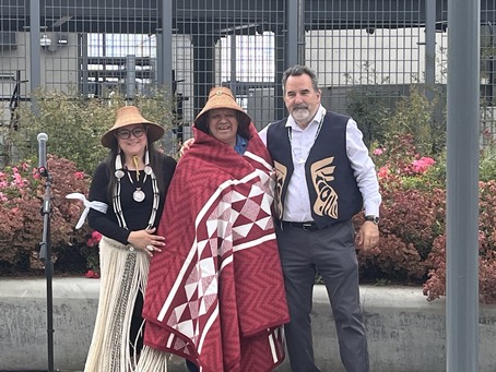 Three people outdoors with greenery behind them. One is wearing traditional attire and a blanket, while another is in a tribal vest.
