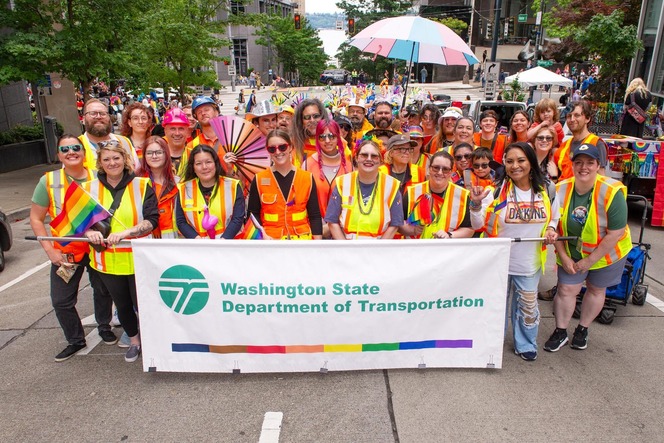A group of people in bright yellow safety vests, holding a banner for the Washington State Department of Transportation at a parade.
