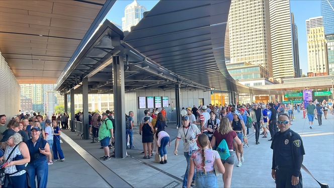 A crowded outdoor area at Seattle's Colman Dock with a long queue of people under a canopy and a security guard nearby.