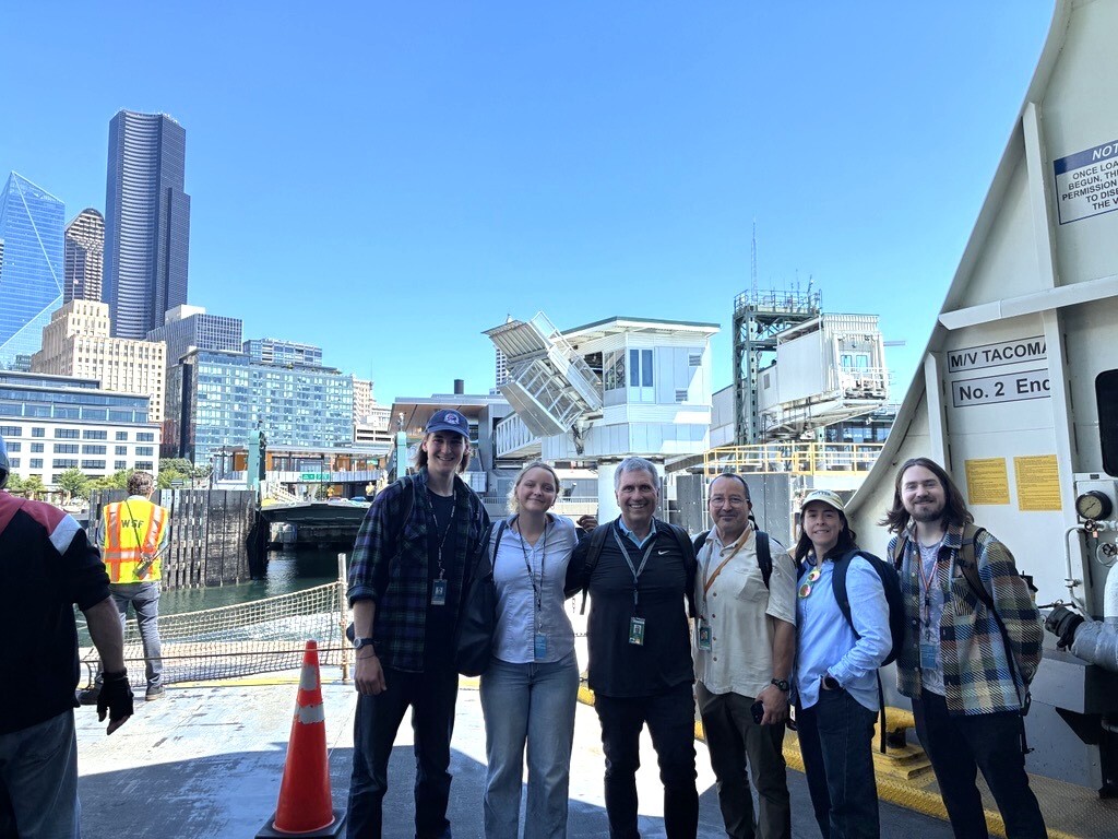 A group of six people on the car deck of a ferry approaching Colman Dock in Seattle with a blue sky and buildings in the background.