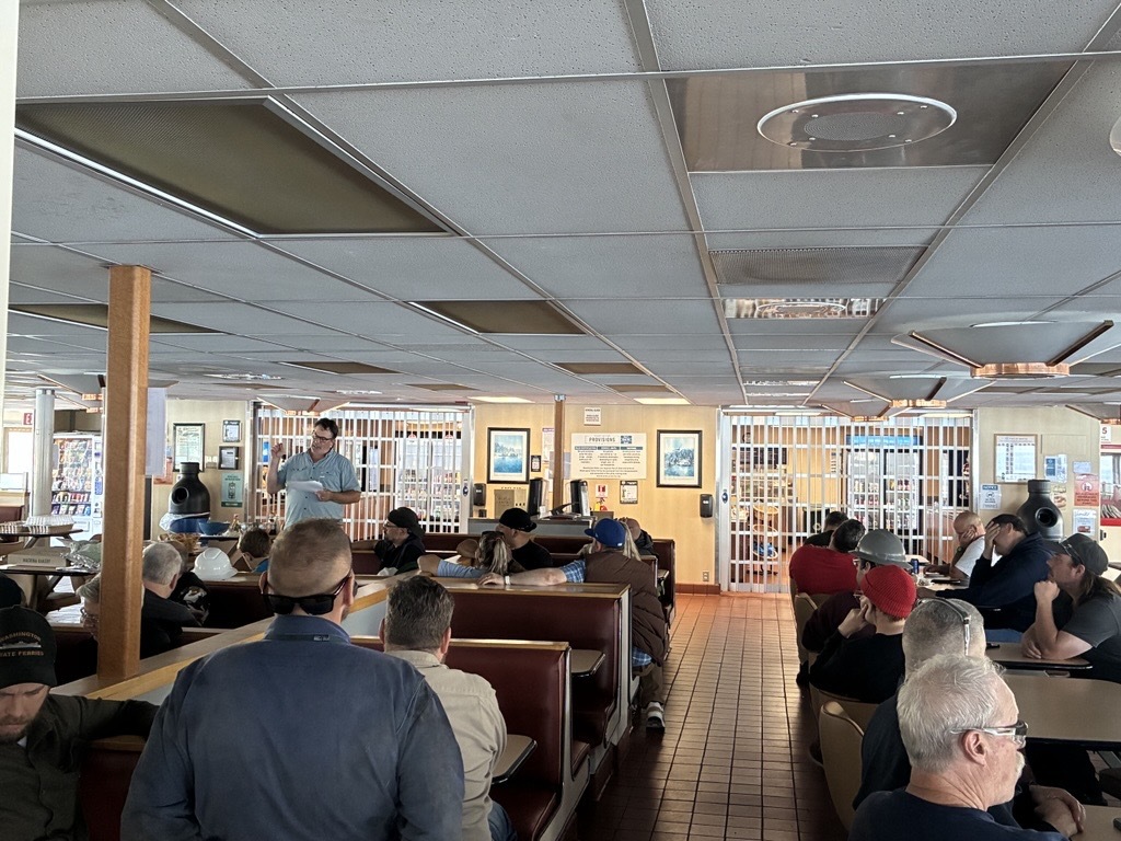 Interior of a ferry with people seated at booths listening to a speaker, surrounded by framed pictures and vending machines.