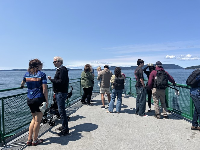 A group of people on an outdoor ferry deck with and distant hills under a clear blue sky.
