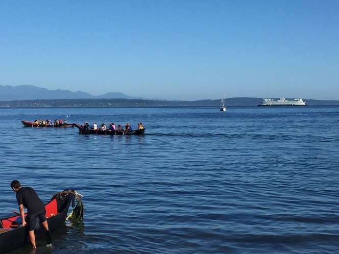 A person adjusts a canoe as groups of people row in the background, with mountains and a ferry under a clear sky.