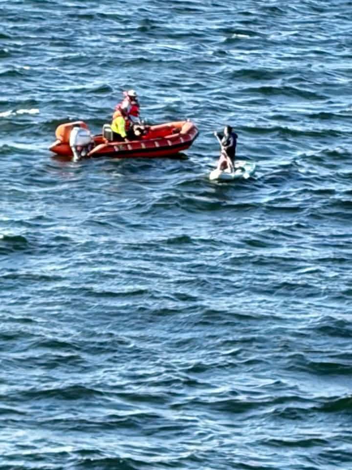 A paddleboarder in a wetsuit navigates choppy blue waters while a small red rescue boat approaches with two individuals in safety gear.