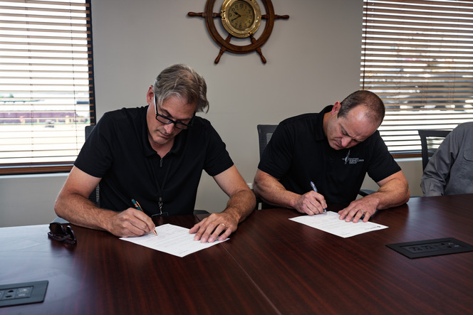Two people signing documents at a conference table in a well-lit room with blinds and a ship's wheel on the wall.
