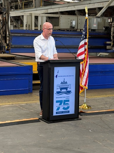 A person speaks at a podium displaying a 75th anniversary Washington State Ferries logo in an industrial setting.