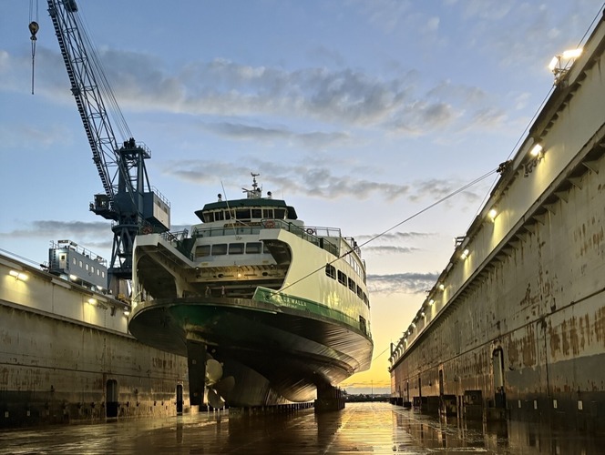 A large green and white ferry is dry-docked in an industrial setting at sunset, with a crane on the left side and reflections in calm water.