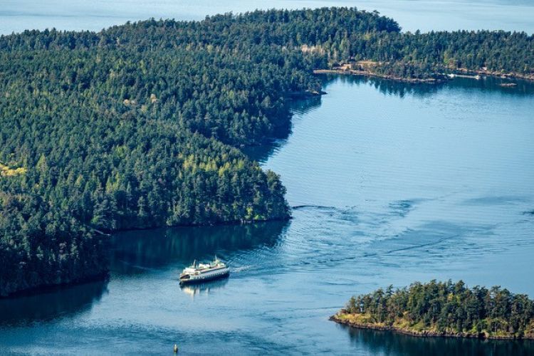 A ferry navigates a winding channel between green forests and islands in a calm water body.