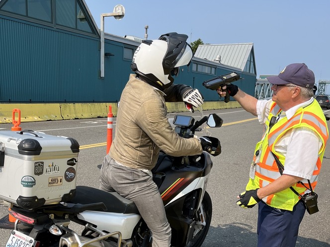 A motorcycle rider is being checked by an Edmonds terminal employee in a bright safety vest on a road with traffic cones.
