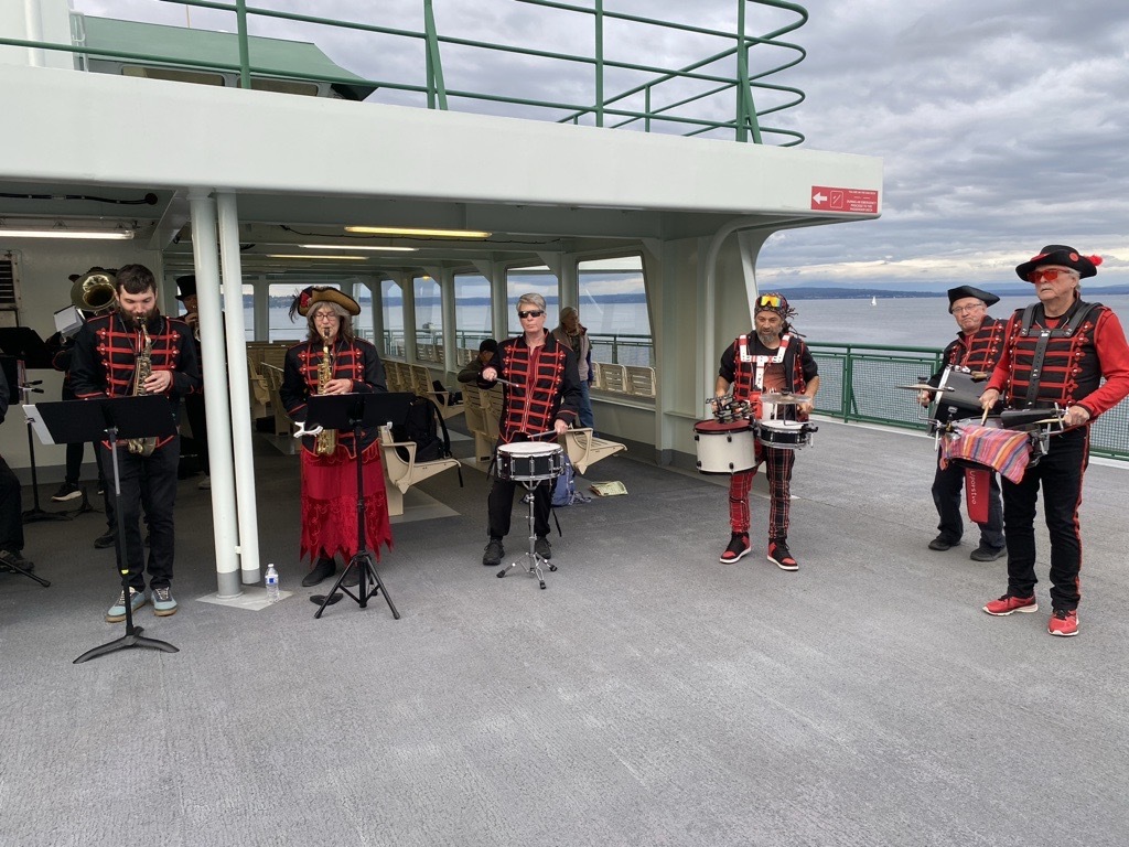 Costumed musicians perform with saxophones and drums on an open ferry deck overlooking the water.