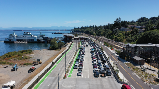 Aerial view of a Mukilteo terminal with a docked ferry, crowded parking lot, railway tracks, hillside homes and distant mountains.