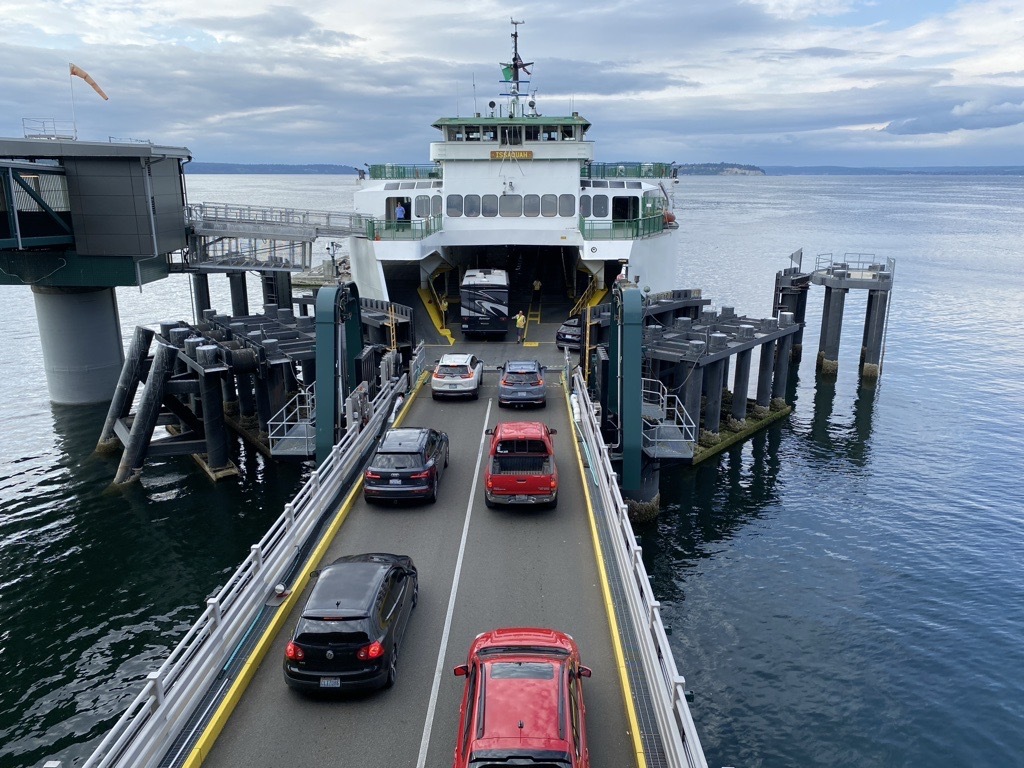 Cars drive up a loading ramp onto a white ferry surrounded by terminal pilings and calm water.