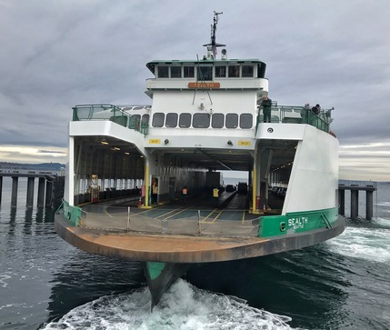 Front view of a white-and-green ferry named Sealth moving away from a dock, with cars visible on its open deck.