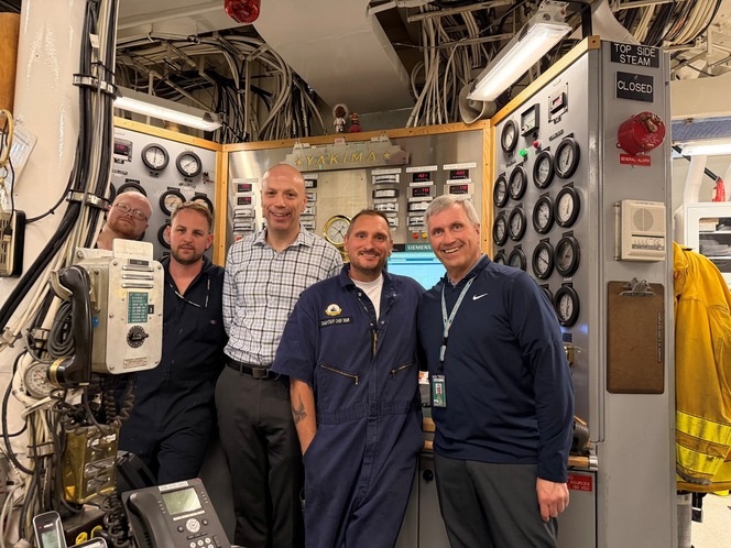 Five people pose in a densely wired ship engine-control room surrounded by gauges and control panels labeled “YAKIMA.”