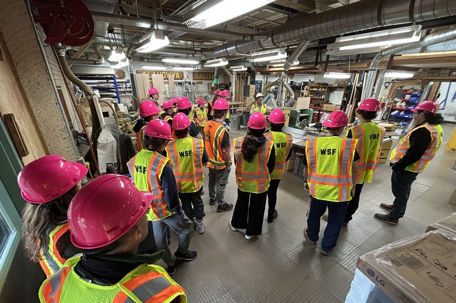 Group wearing pink hard hats and reflective safety vests gathers inside an industrial woodworking shop.