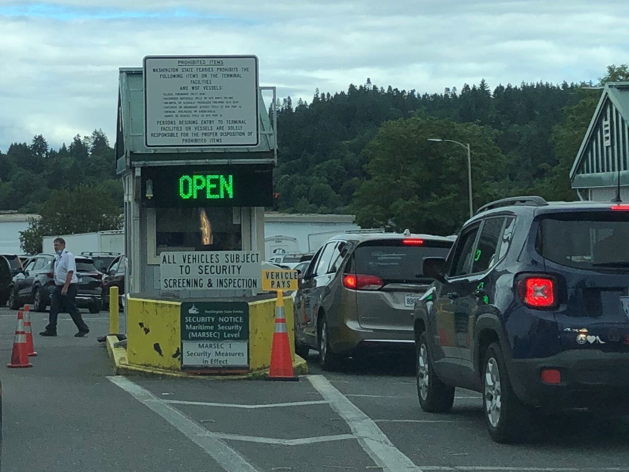Vehicles queue at a Bainbridge terminal toll booth beneath signs about prohibited items and vehicle inspections.