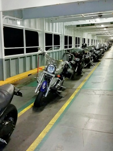 Motorcycles lined up on a ferry's brightly lit enclosed vehicle deck, with an “Engine Off” sign overhead.
