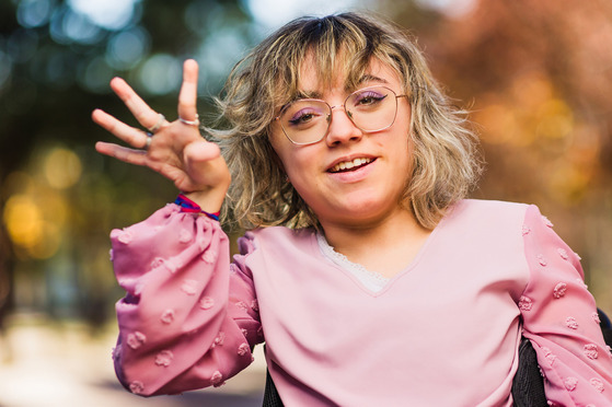 Blonde-haired woman with pink shirt and wire frames waves to the camera