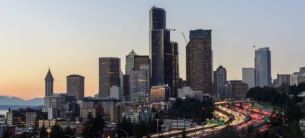 Seattle skyscrapers tower in front of the dim evening sky as cars light up I-5 below them.