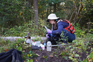 A person crouches on the ground in a forest while filtering water samples into several small and large bottles.
