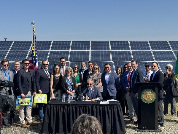 Inslee, legislators and stakeholders smiling at camera after signing of bill, backed by array of solar panels and bright blue sky