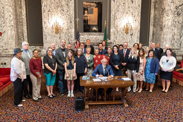 Gov. Jay Inslee is seated at a wooden desk in a large ornate state room surrounded by people awaiting his action on a bill.