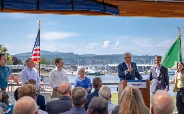 Inslee speaks at podium with other leaders alongside him. Behind him is bright blue sky and picturesque marina.