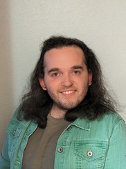 Rueben is smiling & wearing a denim jacket over a knitted tshirt. Rueben’s head is cocked slightly and their shoulder length hair is down.