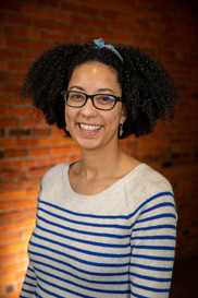 Katherine (Black woman) is pictured in front of a brick wall, smiling. She has an afro, a striped sweater, black glasses, and a blue bow in her hair.
