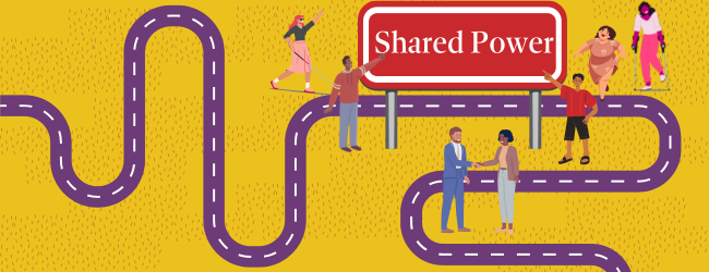 “Shared Power” sits on a red road sign in the middle of a field surrounded by a purple road. A group of people mingle around the sign, joyful.
