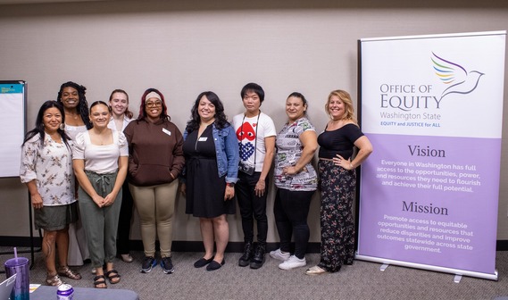 A group of diverse people stand against a beige wall smiling for the camera. They are standing next to an Office of Equity Banner.