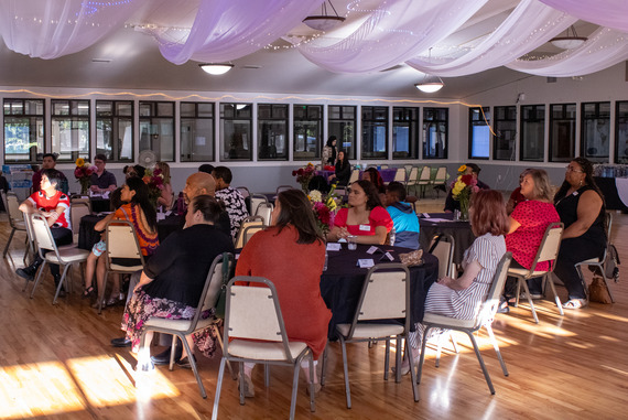 A large group of diverse individuals are seated around tables listening to a presentation. The room is decorated and there are flowers on the tables.