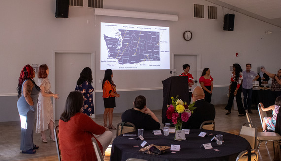 A group of diverse people stand in front of a seated group. Behind them is a breakdown of Washington State's counties. 