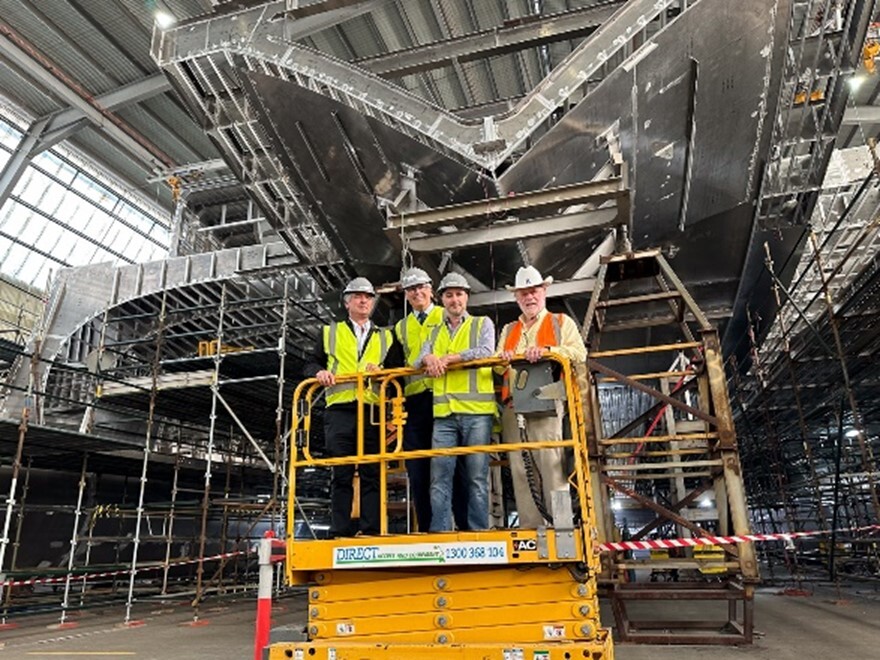 Four men stand on a lift underneath the hull of a large vessel under construction at a shipyard.