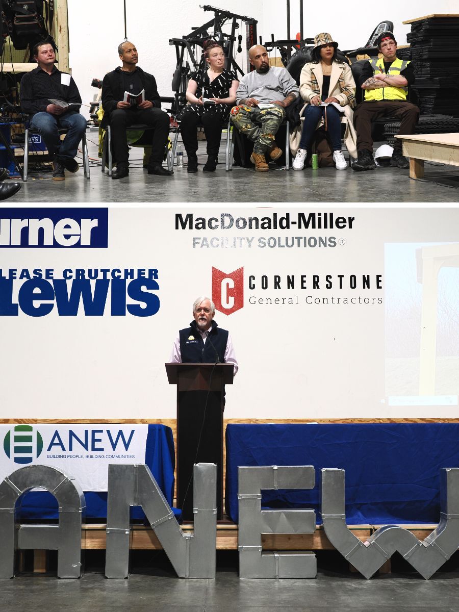 A seated group of people listens to a man deliver a speech in a warehouse setting.