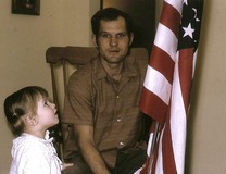 Heidi Helsley as a young child standing next to her father looking at an American flag