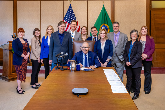 A group of people stand smiling around a seated person who is signing a document.