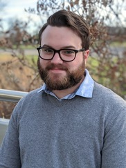 A man from the chest up wearing glasses with brown hair, a mustache and beard, and a relaxed smile wearing a sweater over a collared shirt.